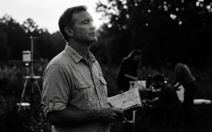 A man with a notebook in a field at dusk while others work behind him, on sunlight as a biological necessity