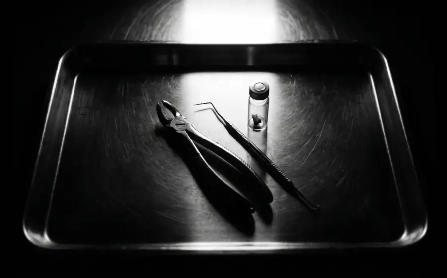 A steel instrument tray holding dental extraction forceps, a probe and a specimen vial, on postmortem pink teeth