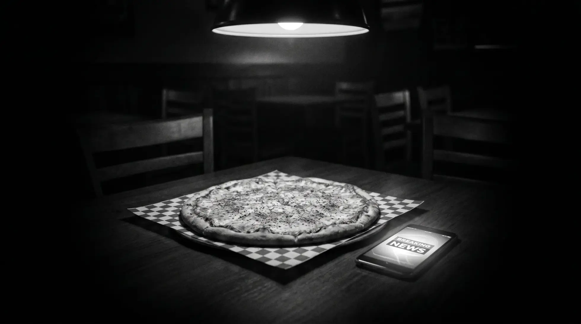 Cold pizza and smartphone with breaking-news alert in a darkened Italian restaurant, black and white.