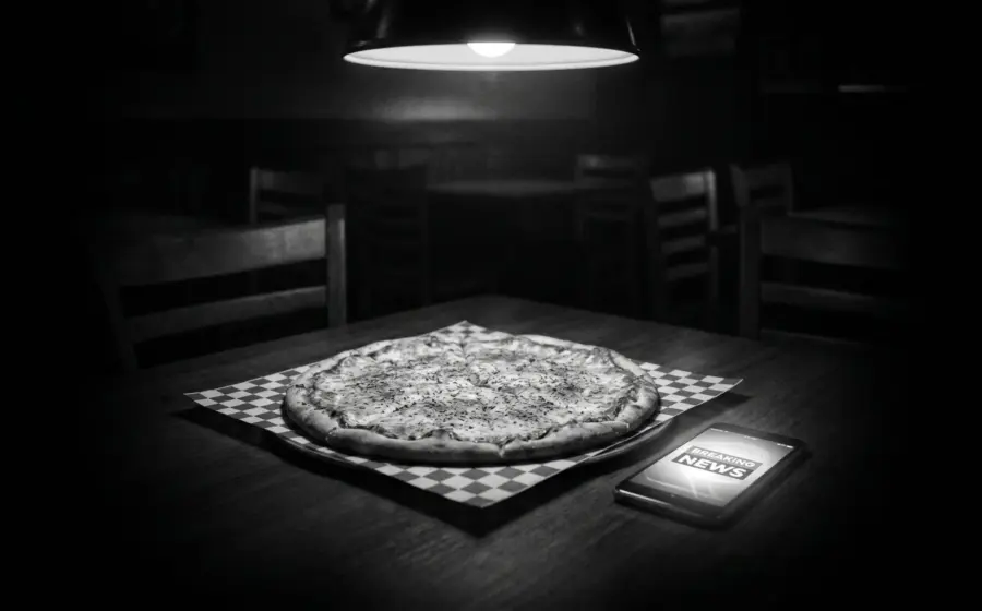 Cold pizza and smartphone with breaking-news alert in a darkened Italian restaurant, black and white.