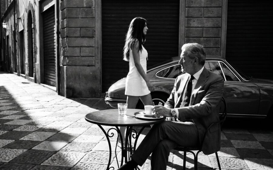 Älterer Mann mit Rolex am Café in italienischer Gasse, Ferrari vorne, junge brunette Frau im weißen Kleid geht vorbei, s/w.
