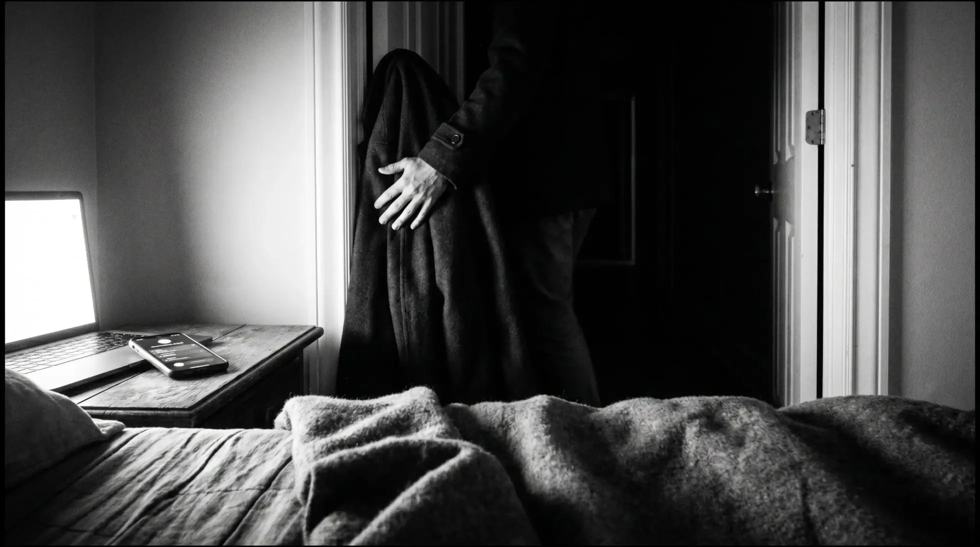 A figure carrying a coat leaves a dim bedroom past a lit laptop and a phone, on walking out of the noise into silence