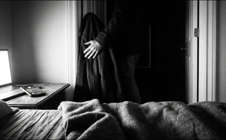 A figure carrying a coat leaves a dim bedroom past a lit laptop and a phone, on walking out of the noise into silence