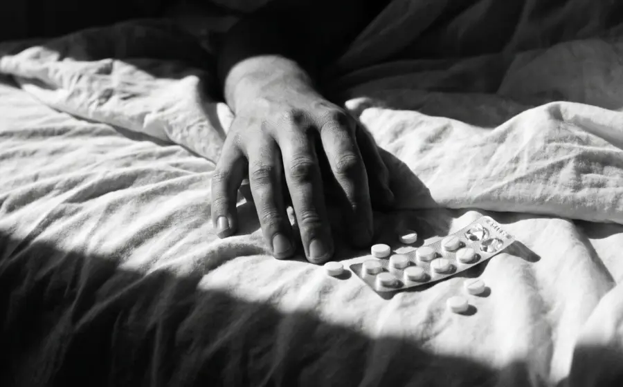 A hand resting on a bed sheet beside loose tablets and an opened blister pack, on the quiet calm that precedes a suicide