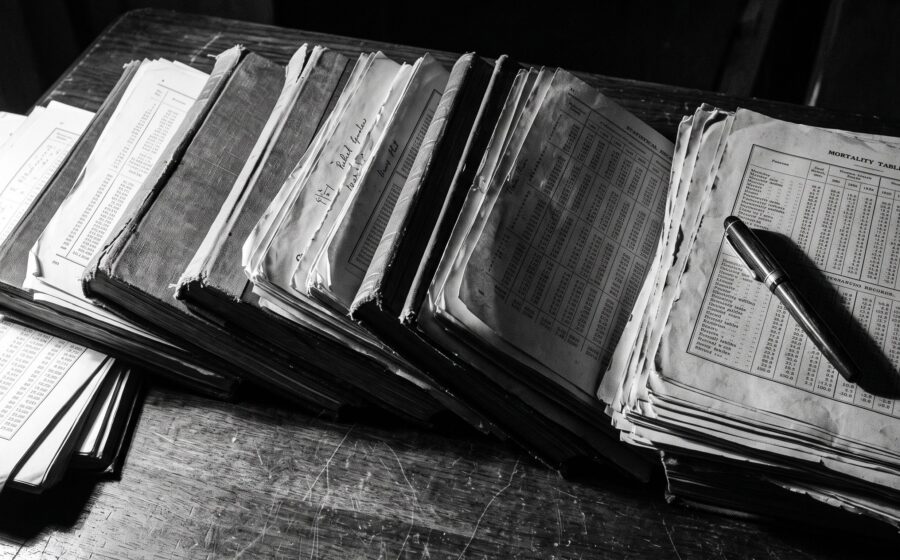 Black and white desk covered edge to edge with stacked mortality volumes and printed death records under harsh side light.