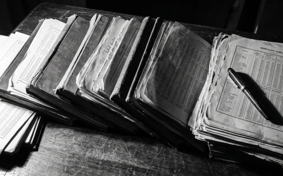 Black and white desk covered edge to edge with stacked mortality volumes and printed death records under harsh side light.