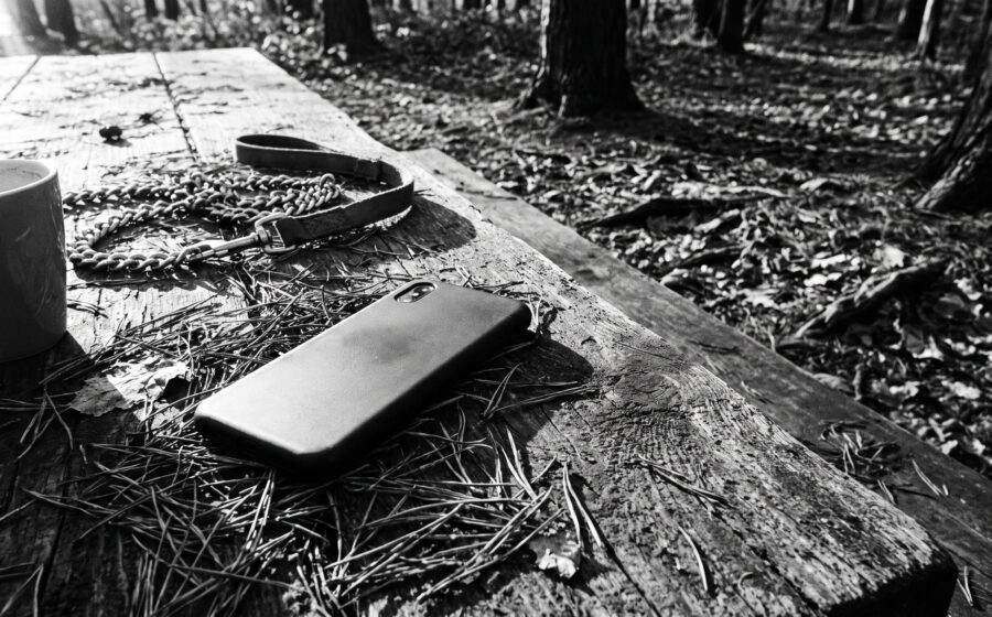 Abandoned face-down smartphone on a wooden forest-edge table beside a dog leash and coffee cup