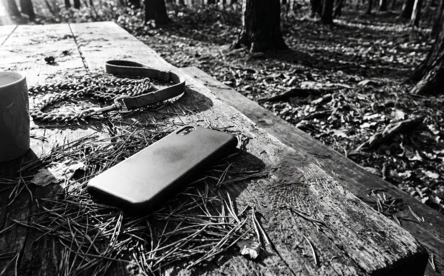 Abandoned face-down smartphone on a wooden forest-edge table beside a dog leash and coffee cup