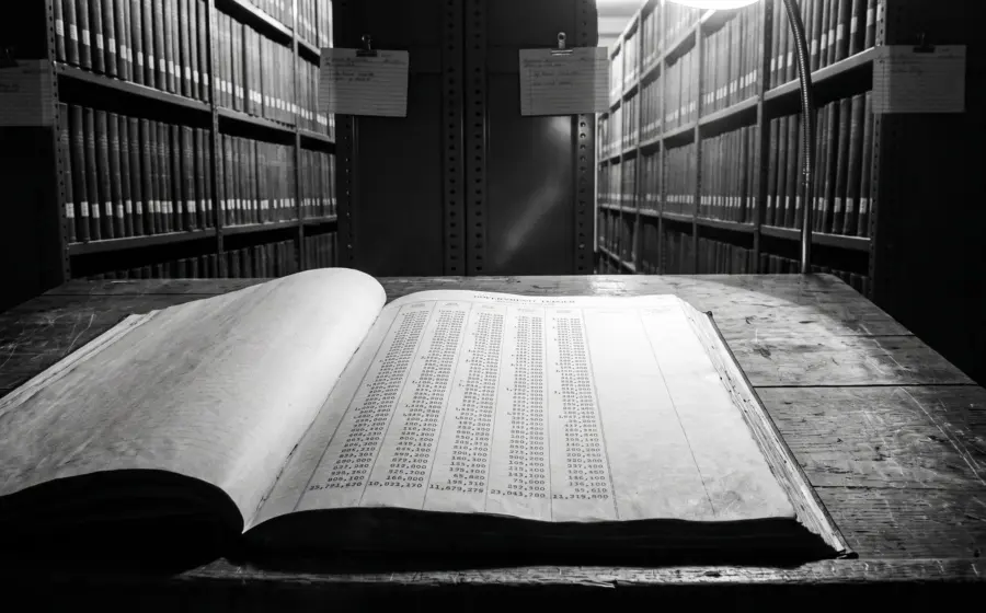 An open government ledger on a worn table in a records hall, its final column left empty, shelves of bound volumes behind